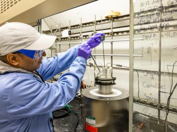 Researcher in a blue coat and glasses, purple gloves and white baseball gat pulls out materials from a metal canister